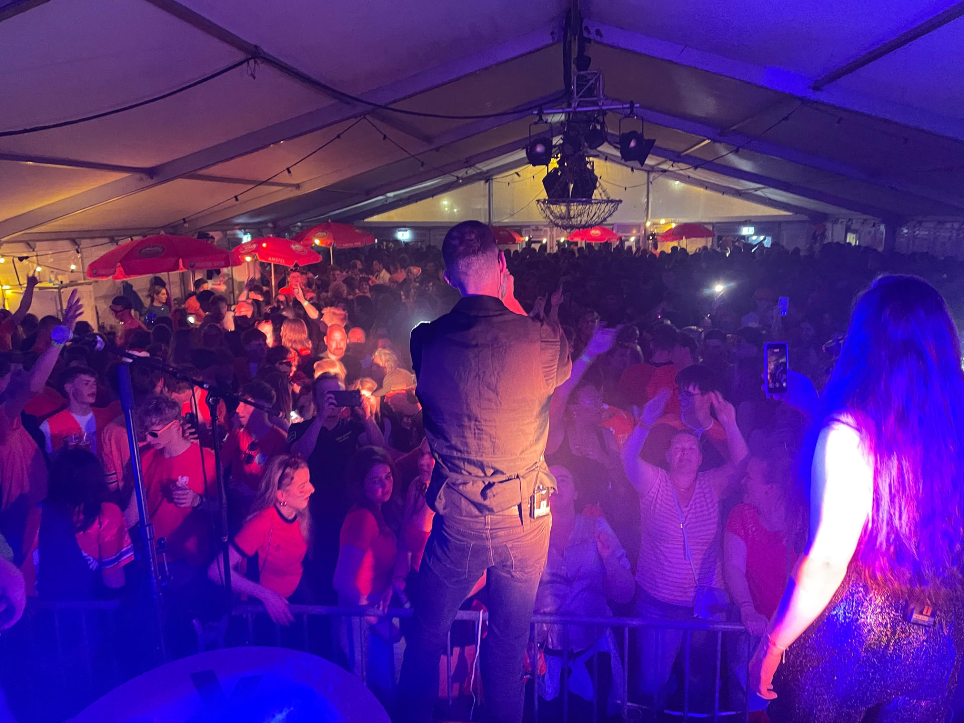 The Hits speelt op het podium van een grote feesttent tijdens Koningsdag; vanaf het podium gezien staat een volle, oranje geklede menigte onder blauw en paars licht mee te zingen