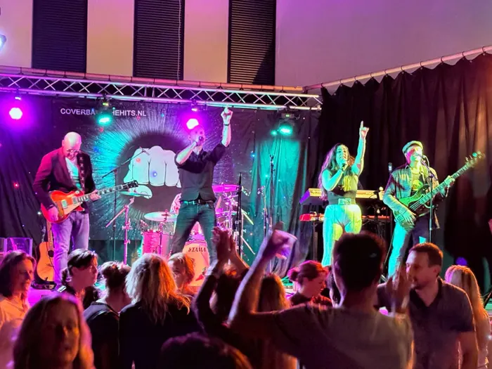 De volledige band van The Hits op het podium met dansend publiek op de voorgrond