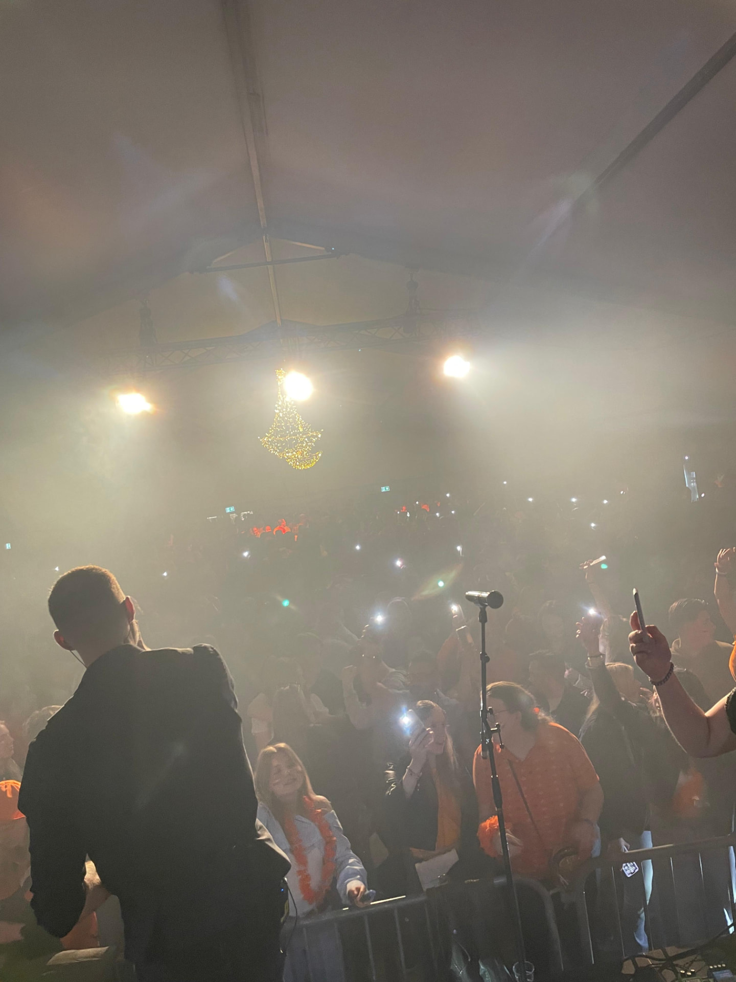 Zanger vanaf het podium gezien in een mistige feesttent, met voor hem een volle zaal die smartphones met zaklampen omhoog houdt en bezoekers in oranje feestkleding