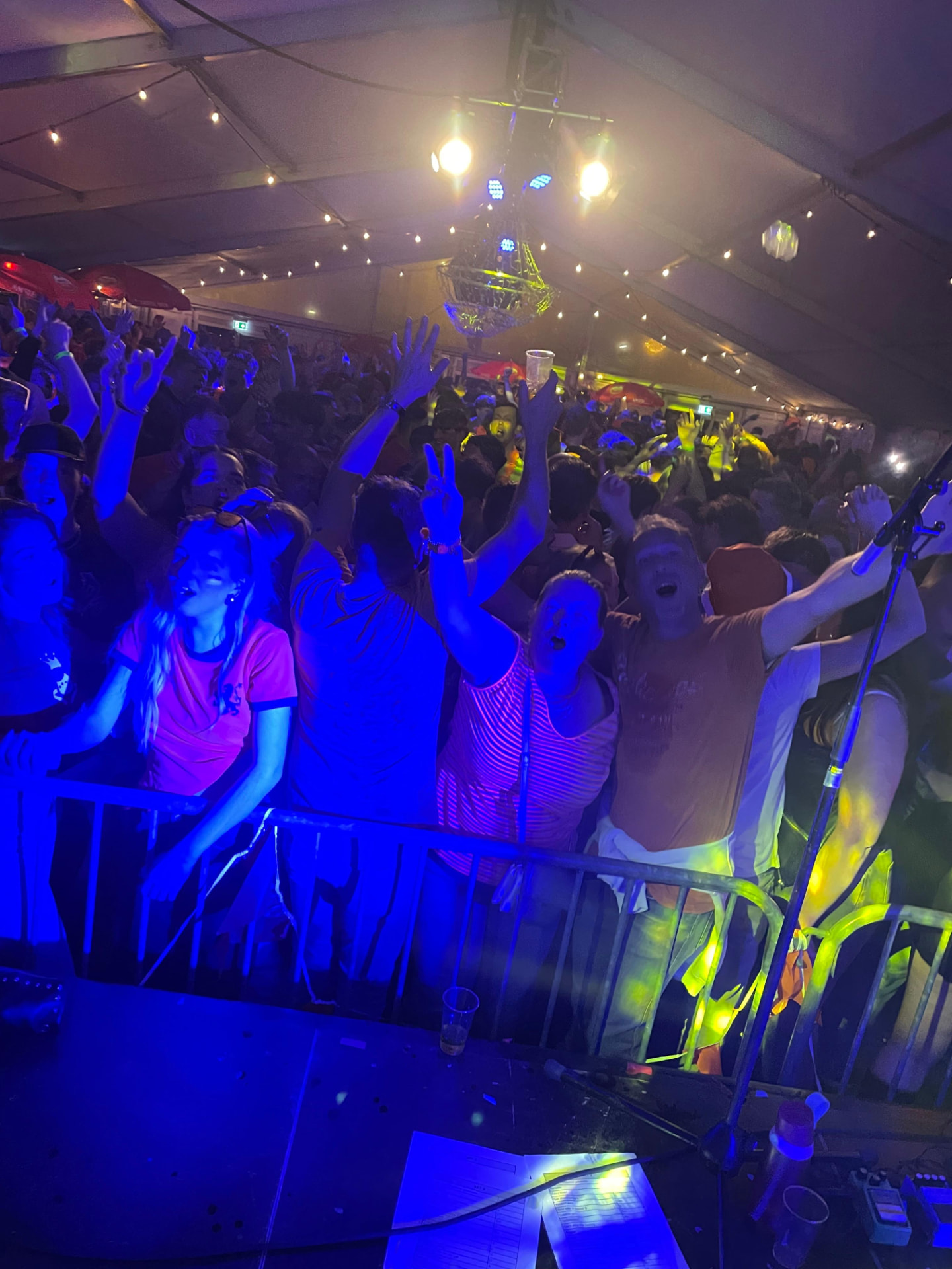 Blik vanaf de podiumrand op de eerste rij van een Koningsdag-feesttent: bezoekers in oranje shirts, met facepaint en opgestoken handen, juichen en zingen mee achter een dranghek