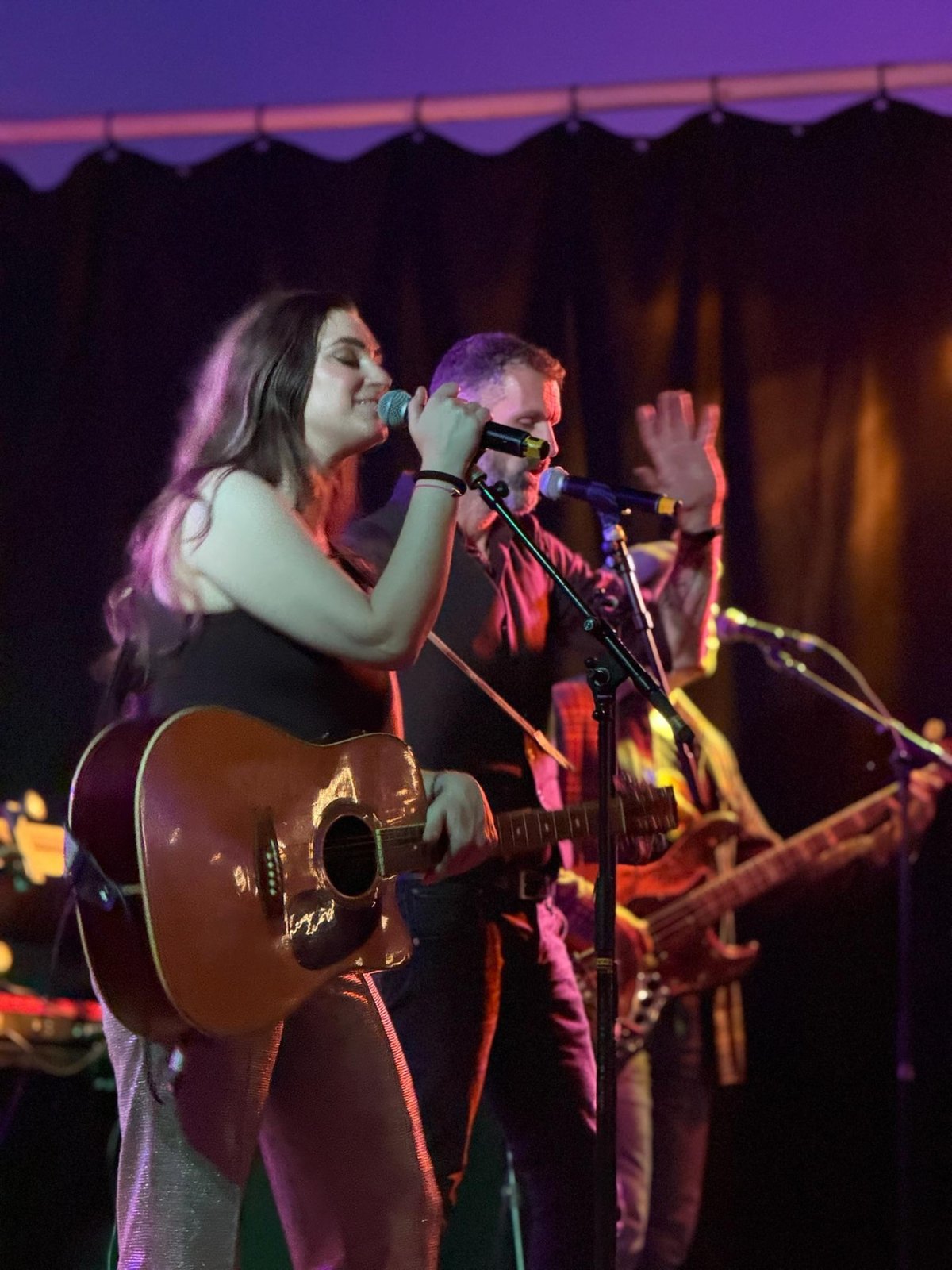 Zangeres van The Hits zingt in de microfoon met een akoestische gitaar, de zanger zingt mee op de achtergrond