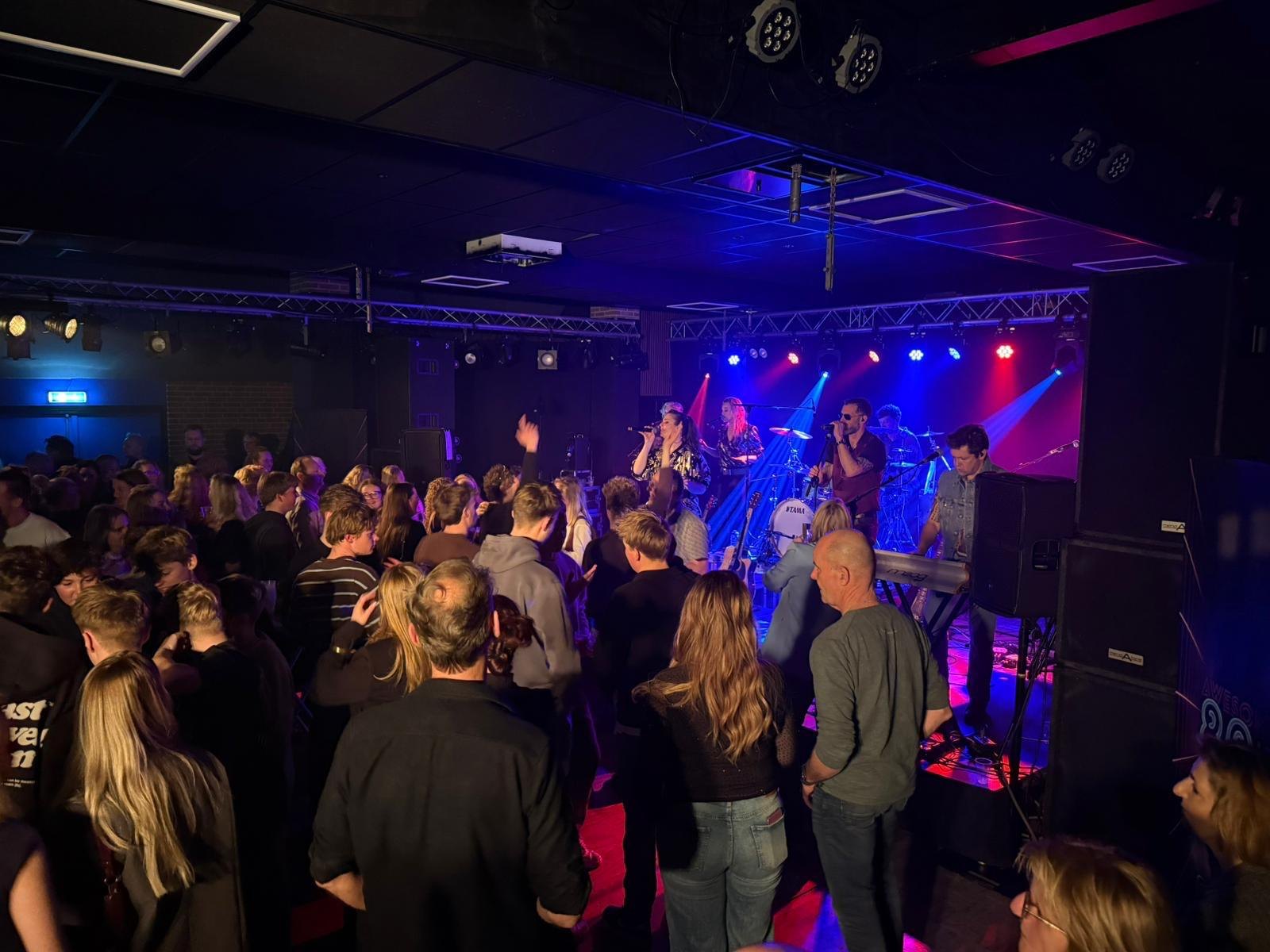 Coverband The Hits speelt op een verlicht podium voor een volle zaal tijdens een feestavond