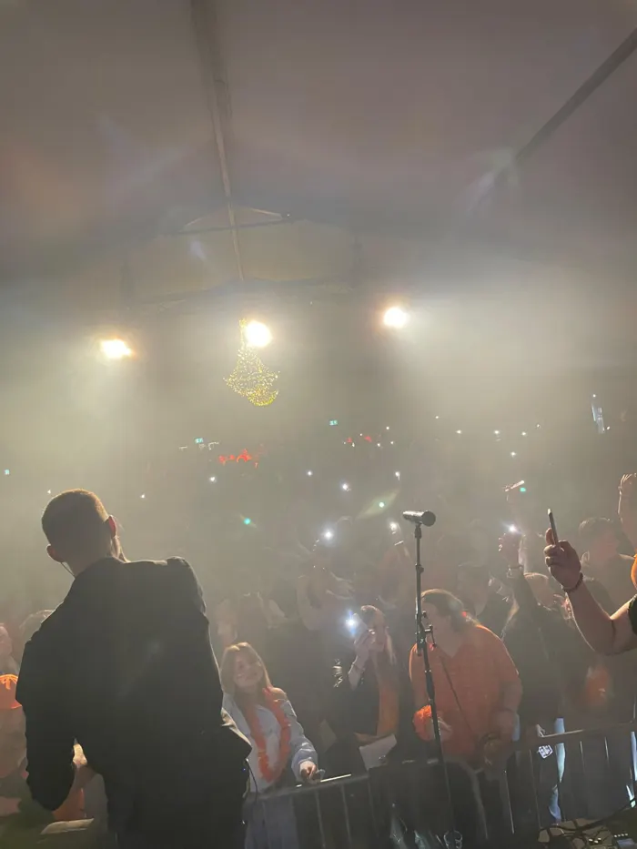 Zanger vanaf het podium gezien in een mistige feesttent, met voor hem een volle zaal die smartphones met zaklampen omhoog houdt en bezoekers in oranje feestkleding