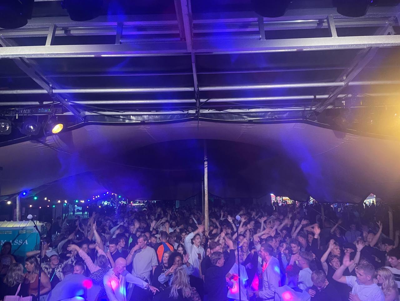 Publiek met handen omhoog onder paars en blauw licht, gezien vanaf het podium van The Hits tijdens een tentfeest