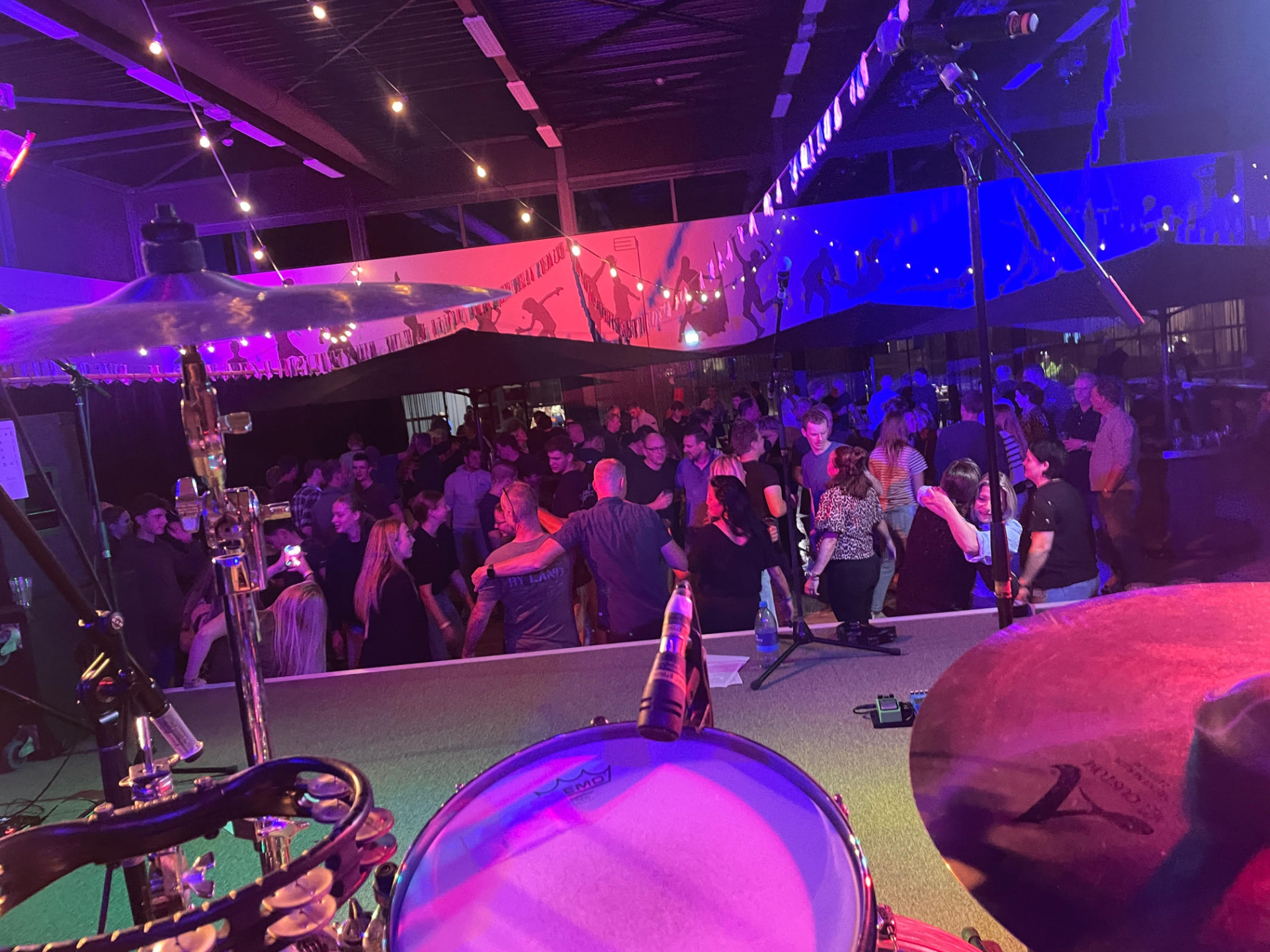 Blik vanaf het podium achter het drumstel van The Hits over een volle, dansende zaal met slingerlampen en paars licht
