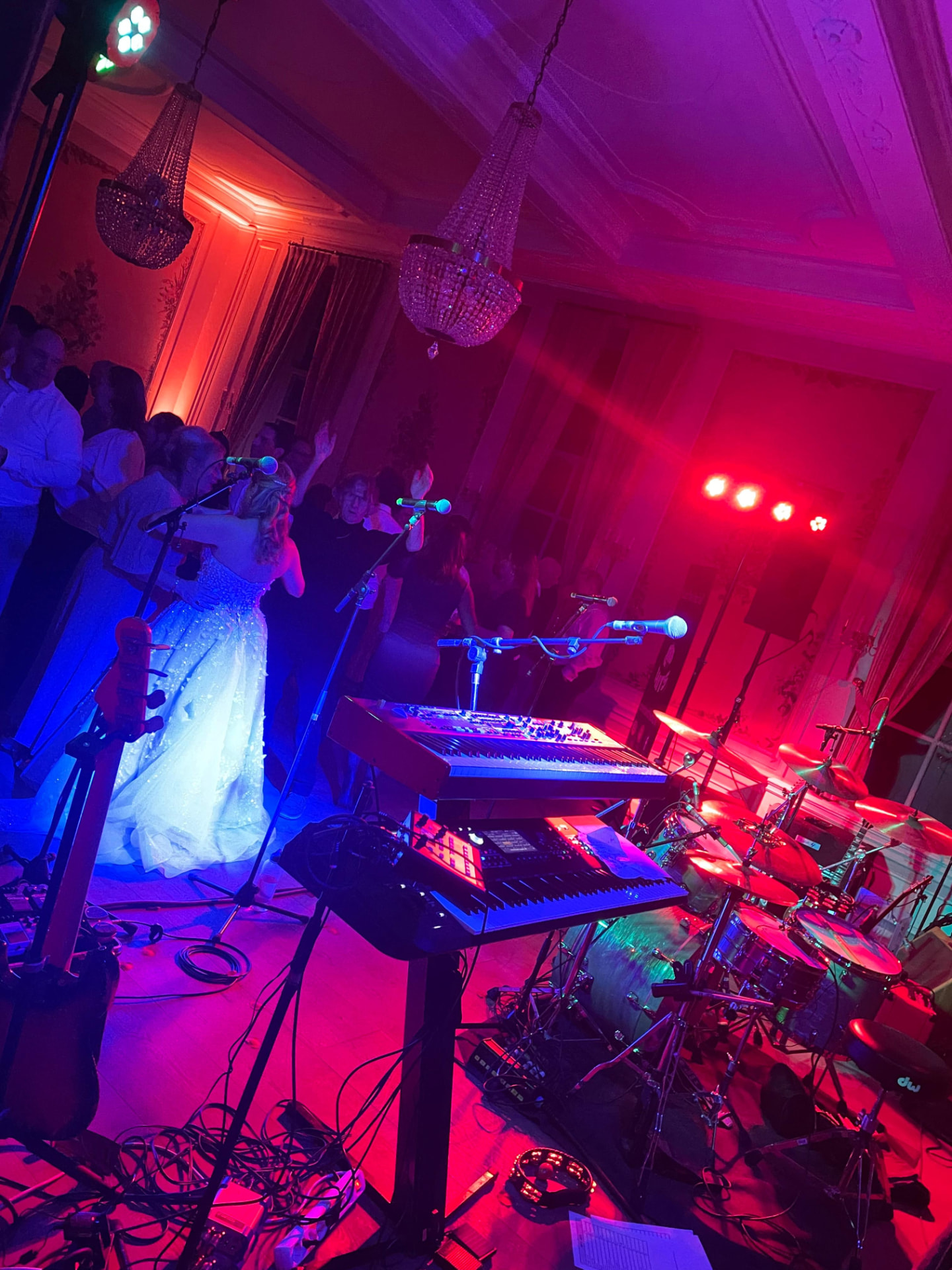 Zicht vanaf het podium met keyboards, drumstel en basgitaar in rood en paars feestlicht, terwijl een bruid en gasten dansen op de dansvloer