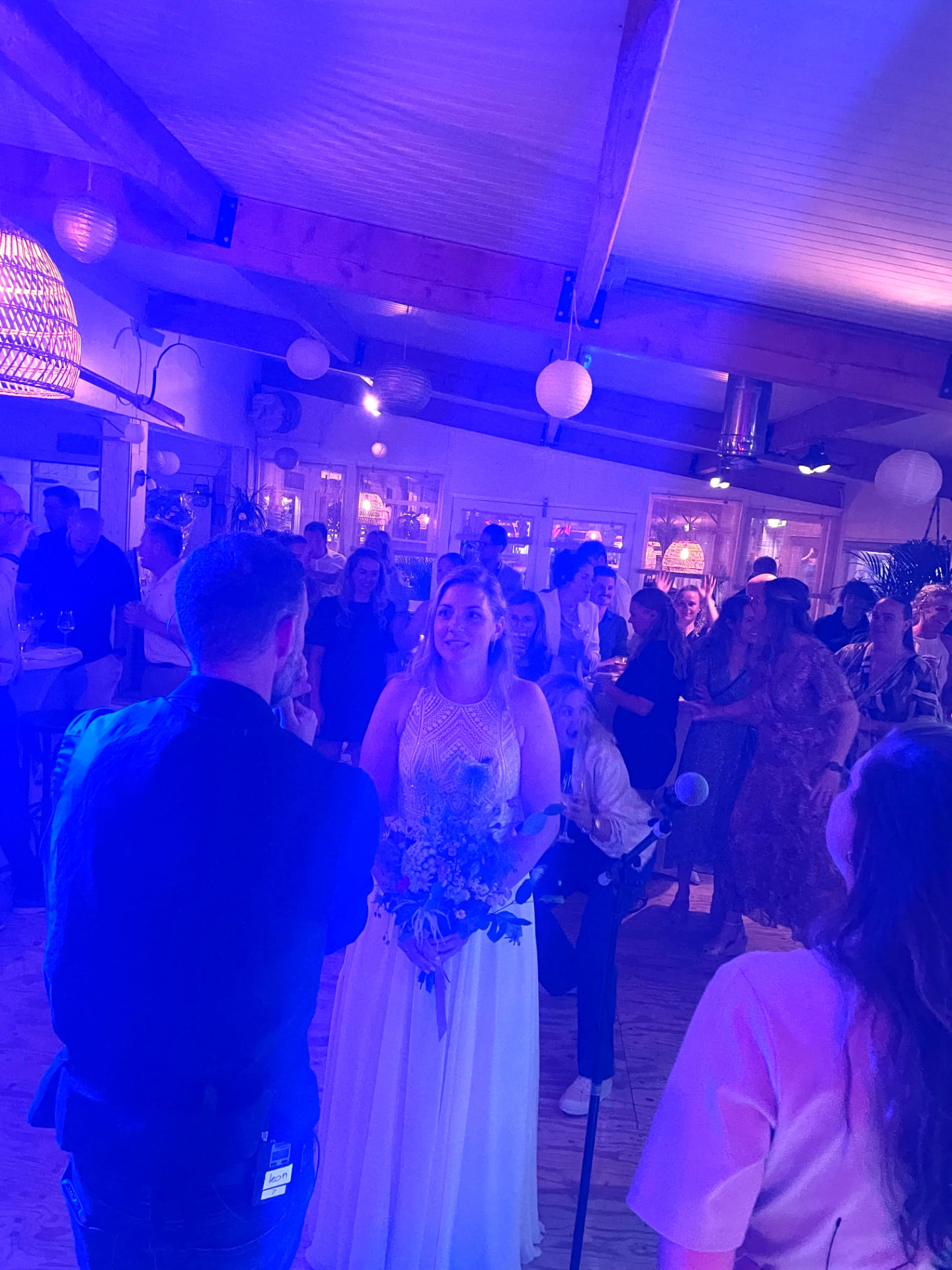 Zanger brengt een serenade aan een glimlachende bruid in een witte trouwjurk met boeket, omringd door feestende gasten in een paarsblauw verlichte zaal